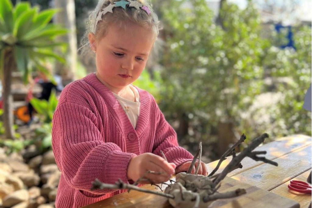 A young child focuses intently on her mud and stick creation, , immersed in creativity and exploration.